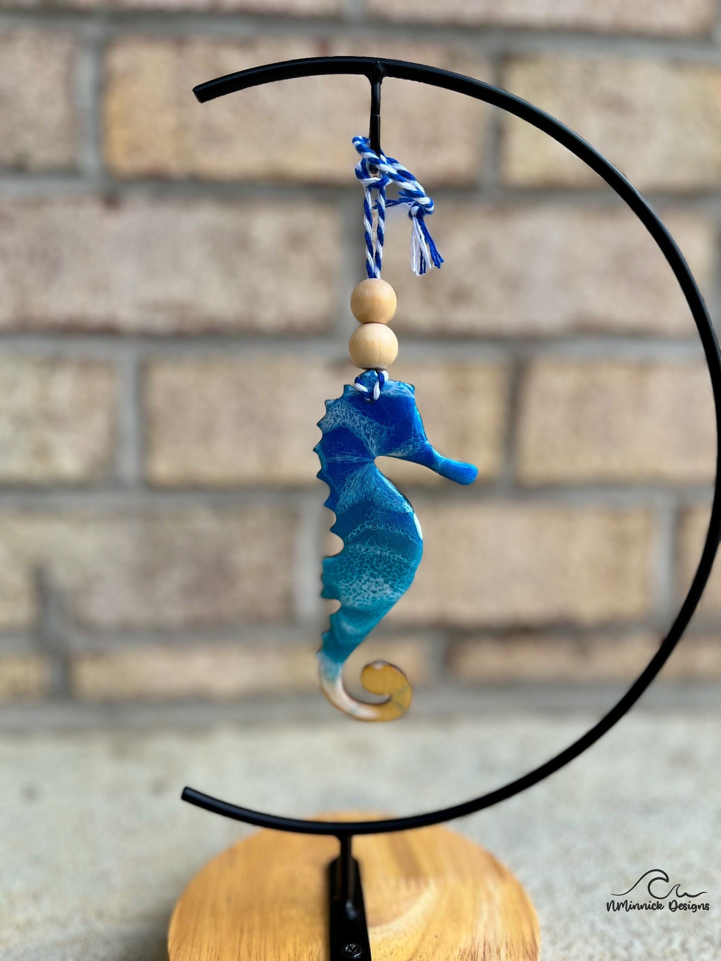 Seahorse shaped wooden ornament with ocean resin waves on the front hanging from blue and white twine with two wooden beads shown on an ornament hanger