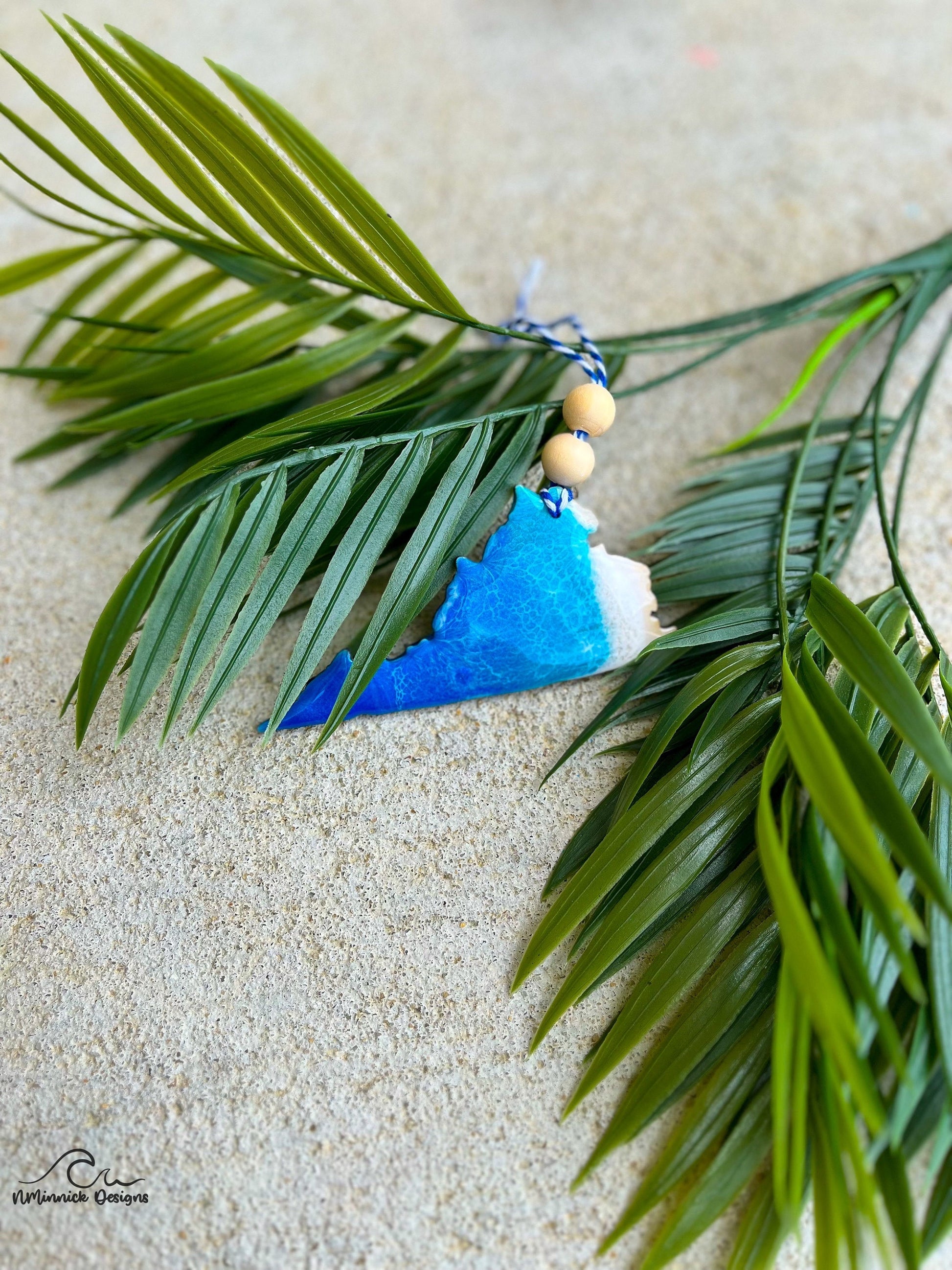 Virginia shaped ornament with ocean resin art laying against palm leaves.