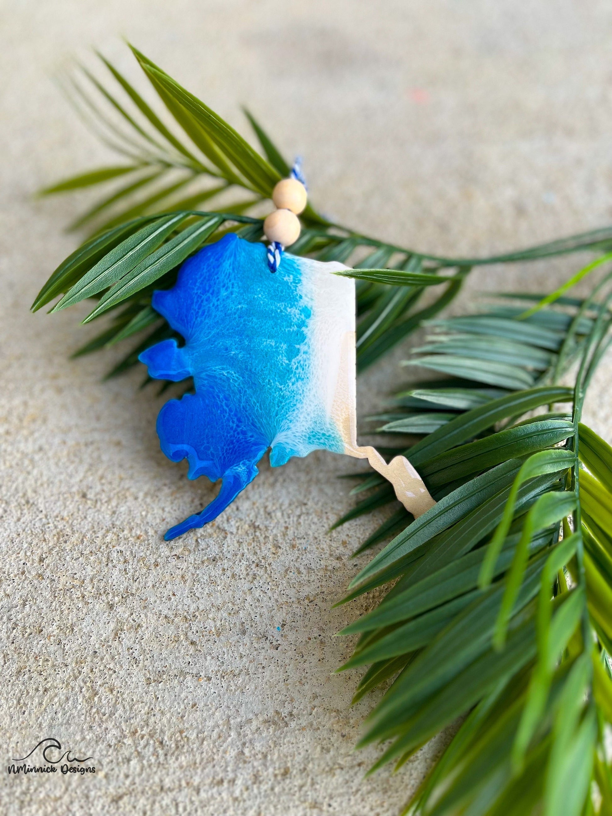 Alaska shaped wooden ornament with ocean resin waves on the front hanging from blue and white twine with two wooden beads and laying against palm leaves