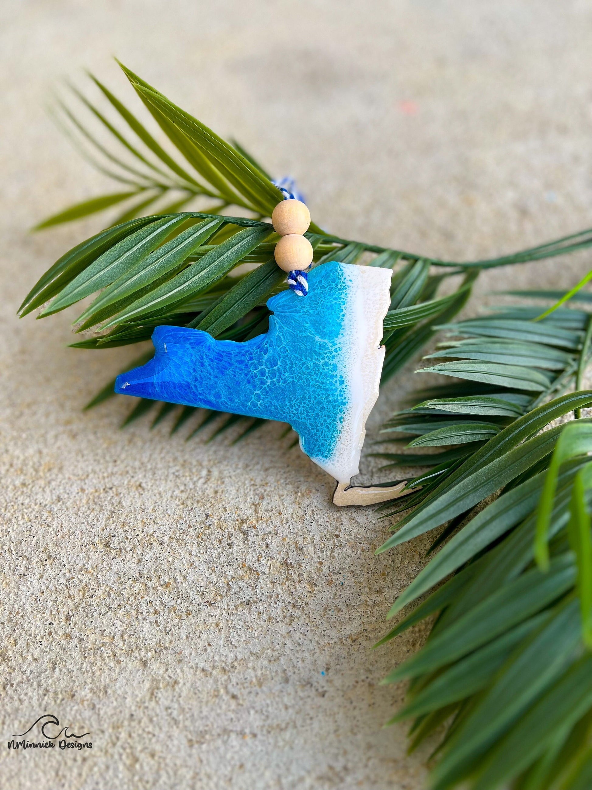 New York shaped wooden ornament with ocean resin waves on the front hanging from blue and white twine with two wooden beads and laying against palm leaves