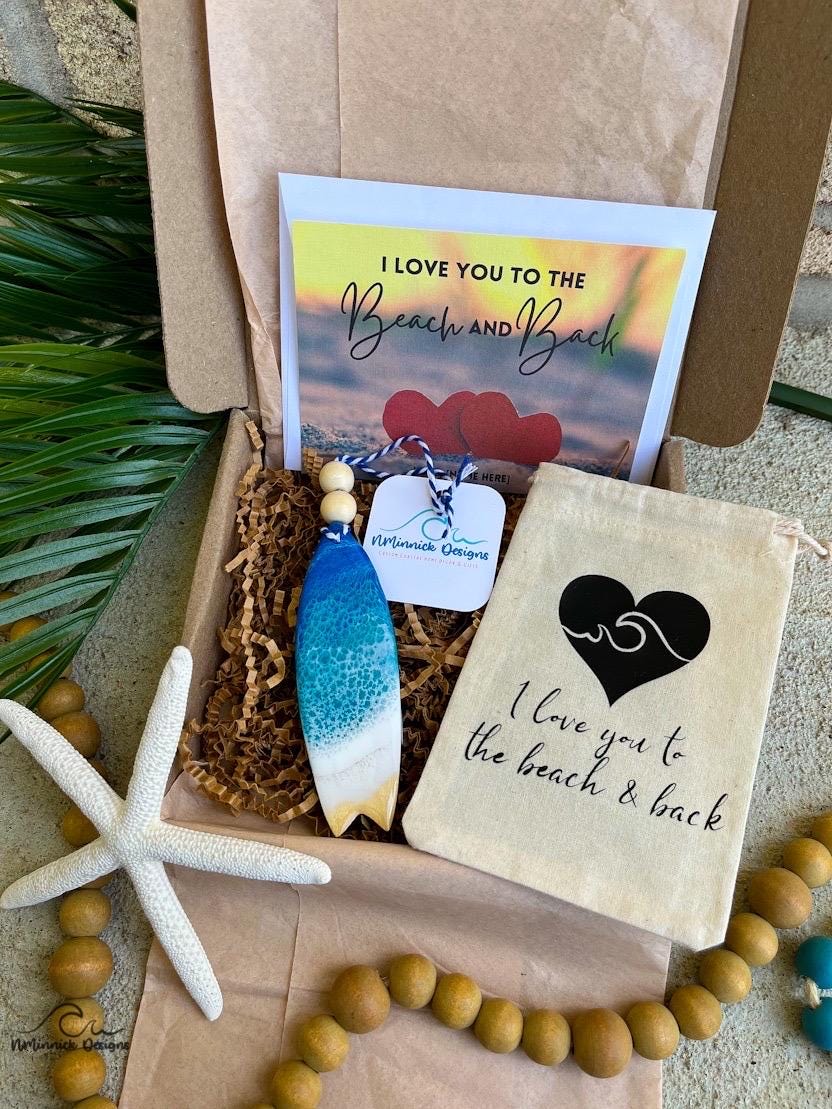 A surfboard shaped wooden hanging ornament with ocean resin art, resembling the waves of the ocean. In a box with an ornament keepsake bag and a custom card.