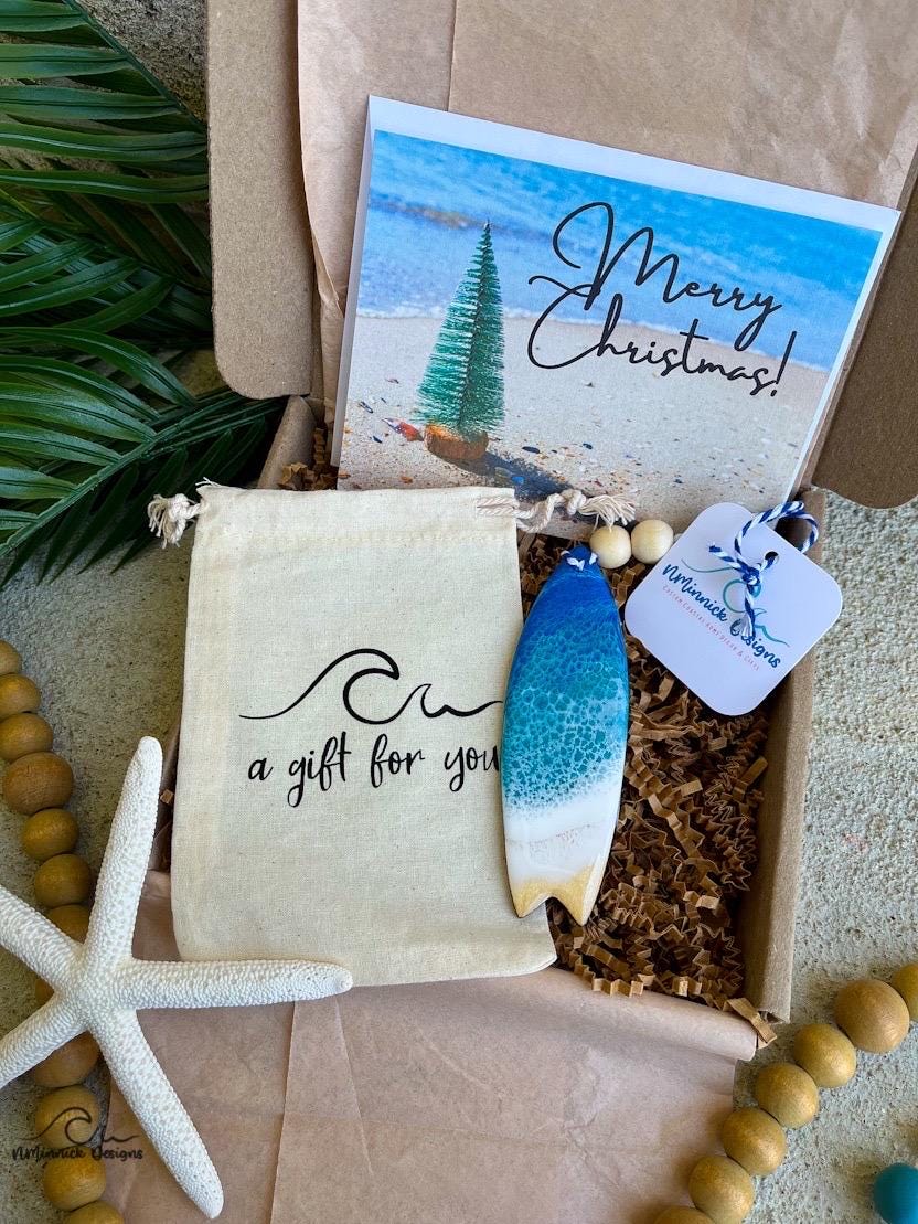 A Surfboard shaped wooden christmas ornament with ocean resin art, resembling the waves of the ocean. In a box with an ornament keepsake bag with a wave that says "a gift for you" and a Merry Christmas custom card.