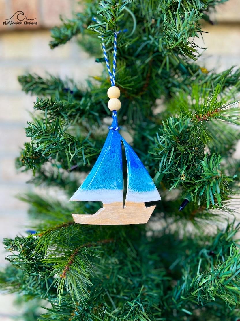 A christmas ornament shaped like a sailboat adorned with a delicate blue and teal ocean resin wave design and hanging on a christmas tree.
