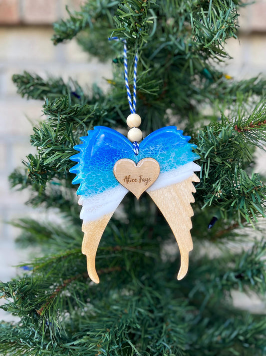 An angel wings memorial keepsake remembrance ornament adorned with ocean resin waves and a heart pendant personalized with the name of a late loved one.