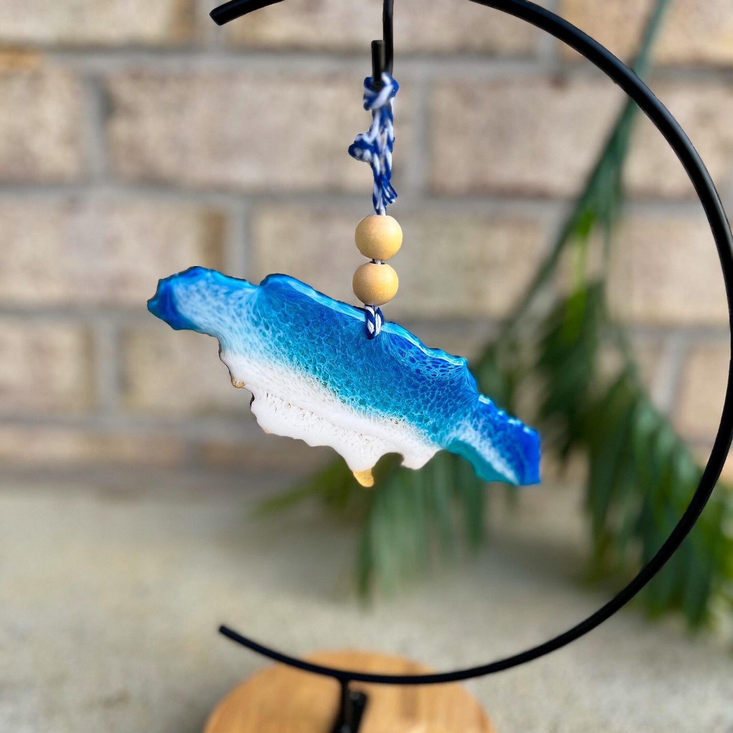 Handmade Jamaica ocean resin ornament featuring blue and teal resin waves over a wooden base shaped like the island of Jamaica, hanging in front of a tropical palm leaf background.