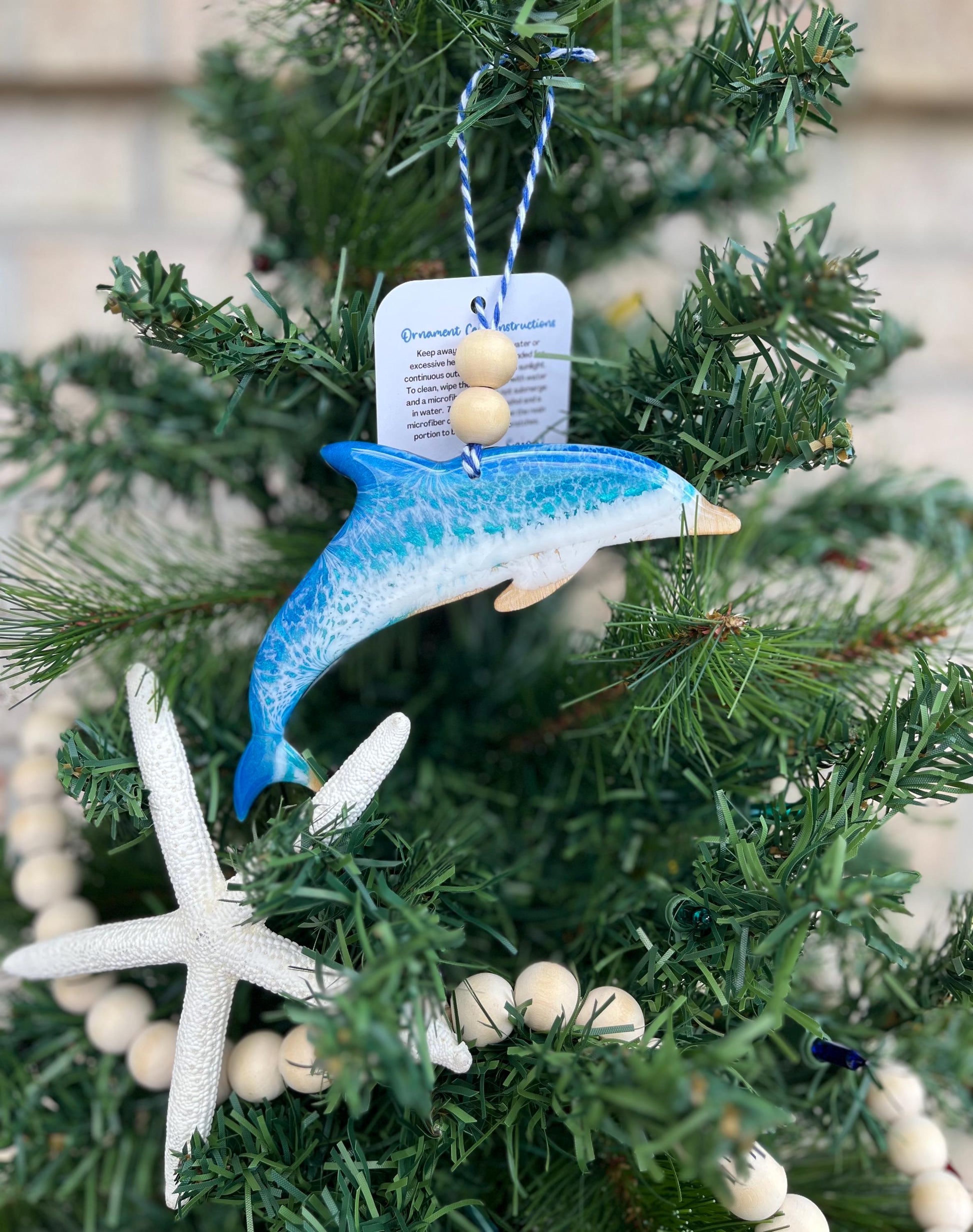 a Dolphin ornament with ocean resin art and finished with two wooden beads and blue and white twine. Hanging on a Christmas tree with a wooden garland and starfish.