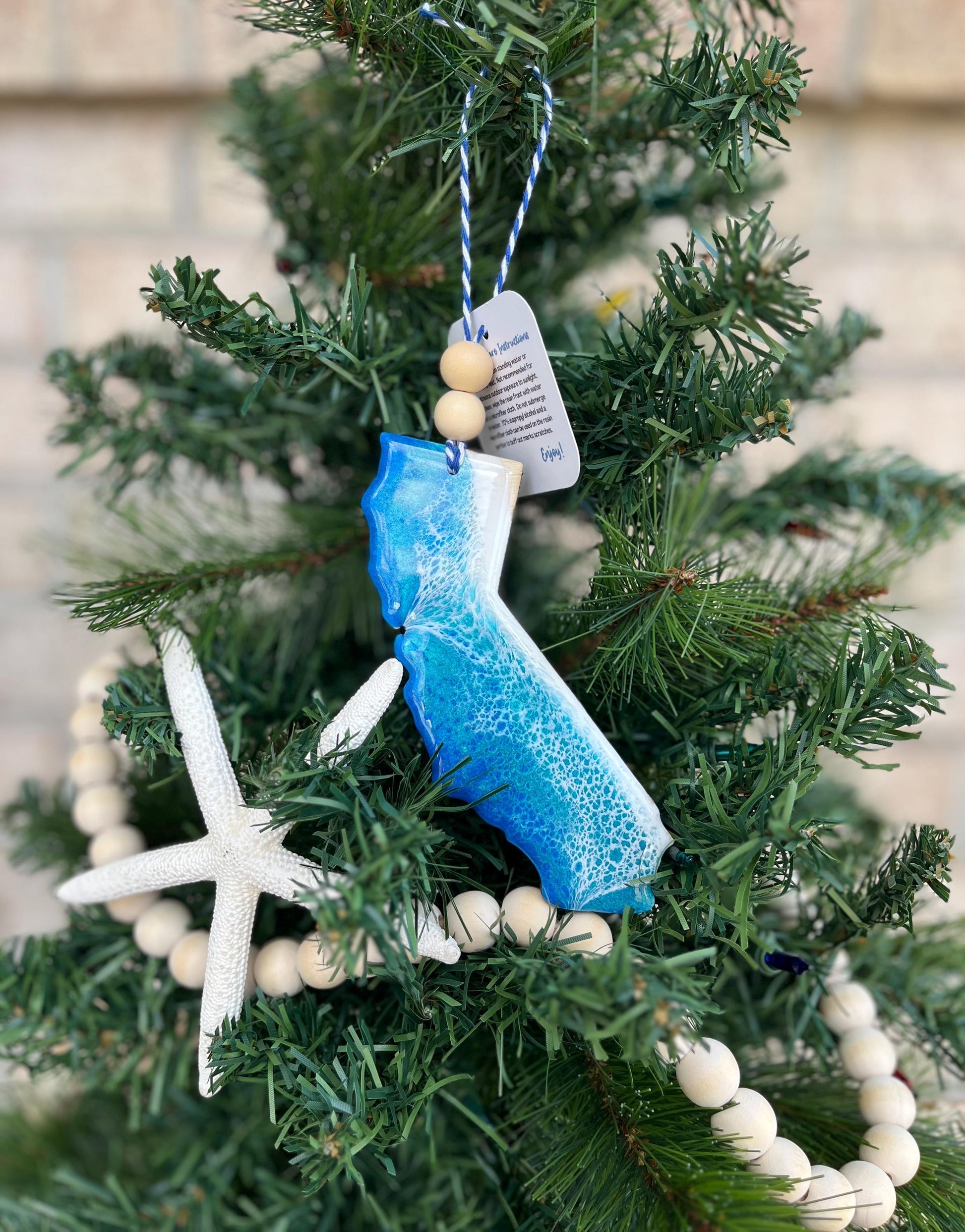 California ornament featuring ocean resin art and finished with two wooden beads and blue and white twine. Hanging on a Christmas tree with a wooden garland and starfish.