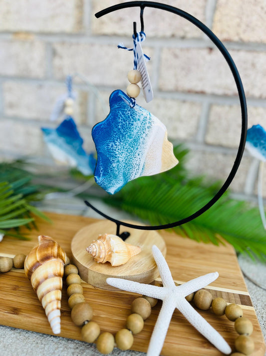 Big Island Hawaii ocean resin ornament hanging on an ornament hanger amongst seashells, palm leaves, and a wooden garland on a bamboo board.