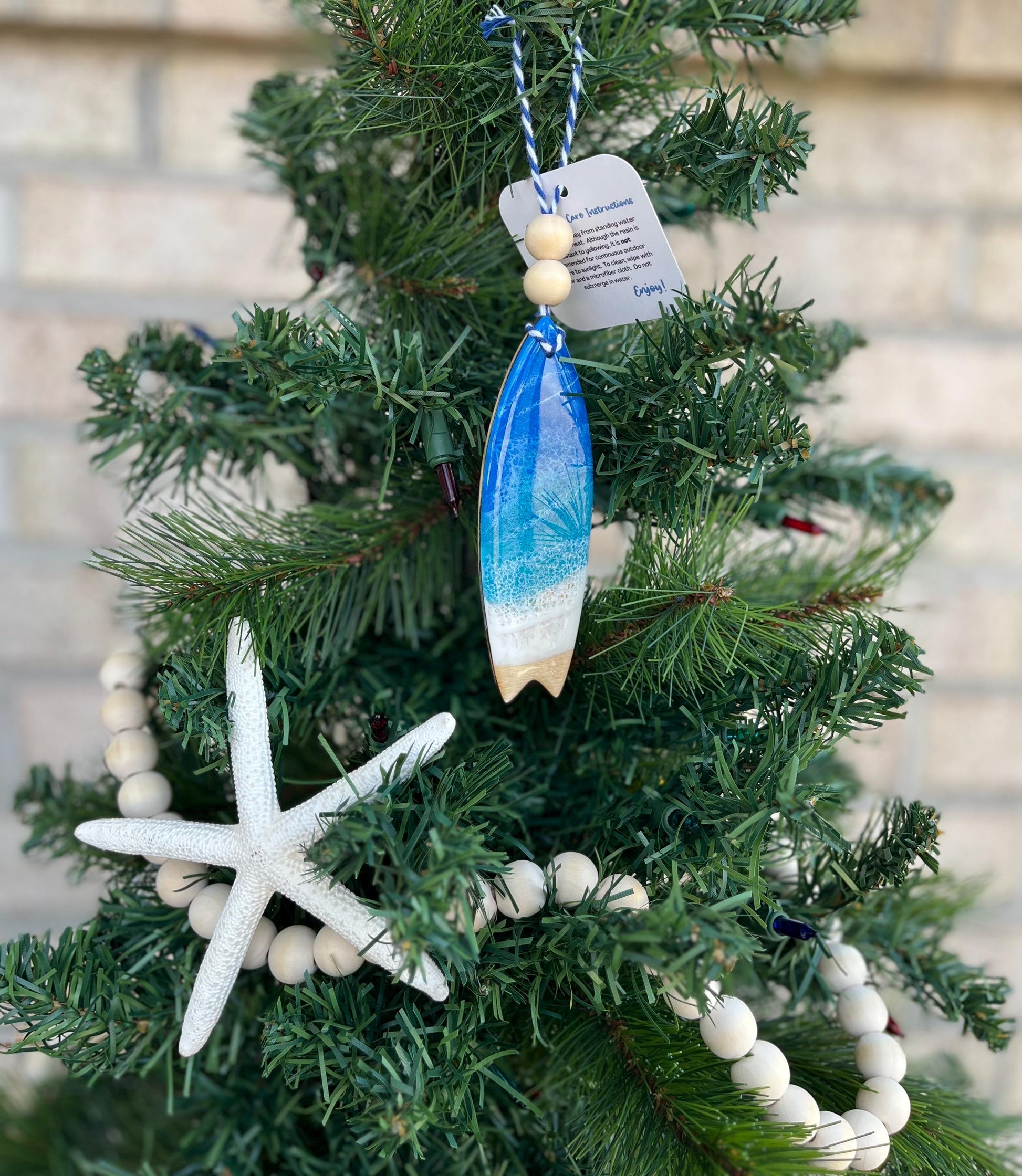 Surfboard ornament featuring ocean resin art and finished with two wooden beads and blue and white twine. Hanging on a Christmas tree with a wooden garland and starfish.