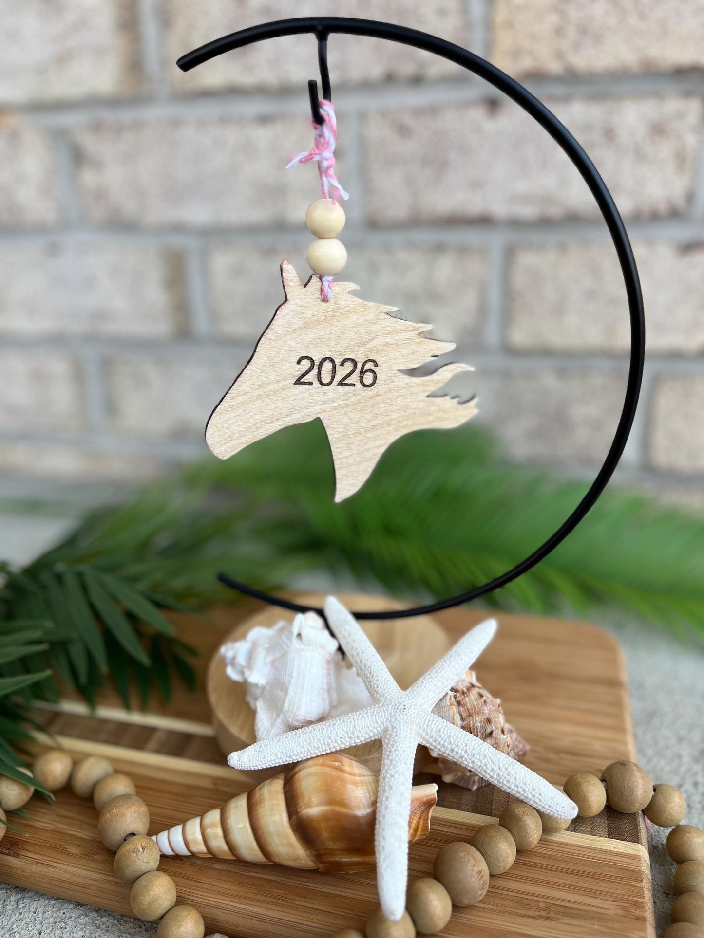 2026 Year of the Horse ornament with 2026 engraved on the back hanging on an ornament hook amongst seashells and palm leaves.
