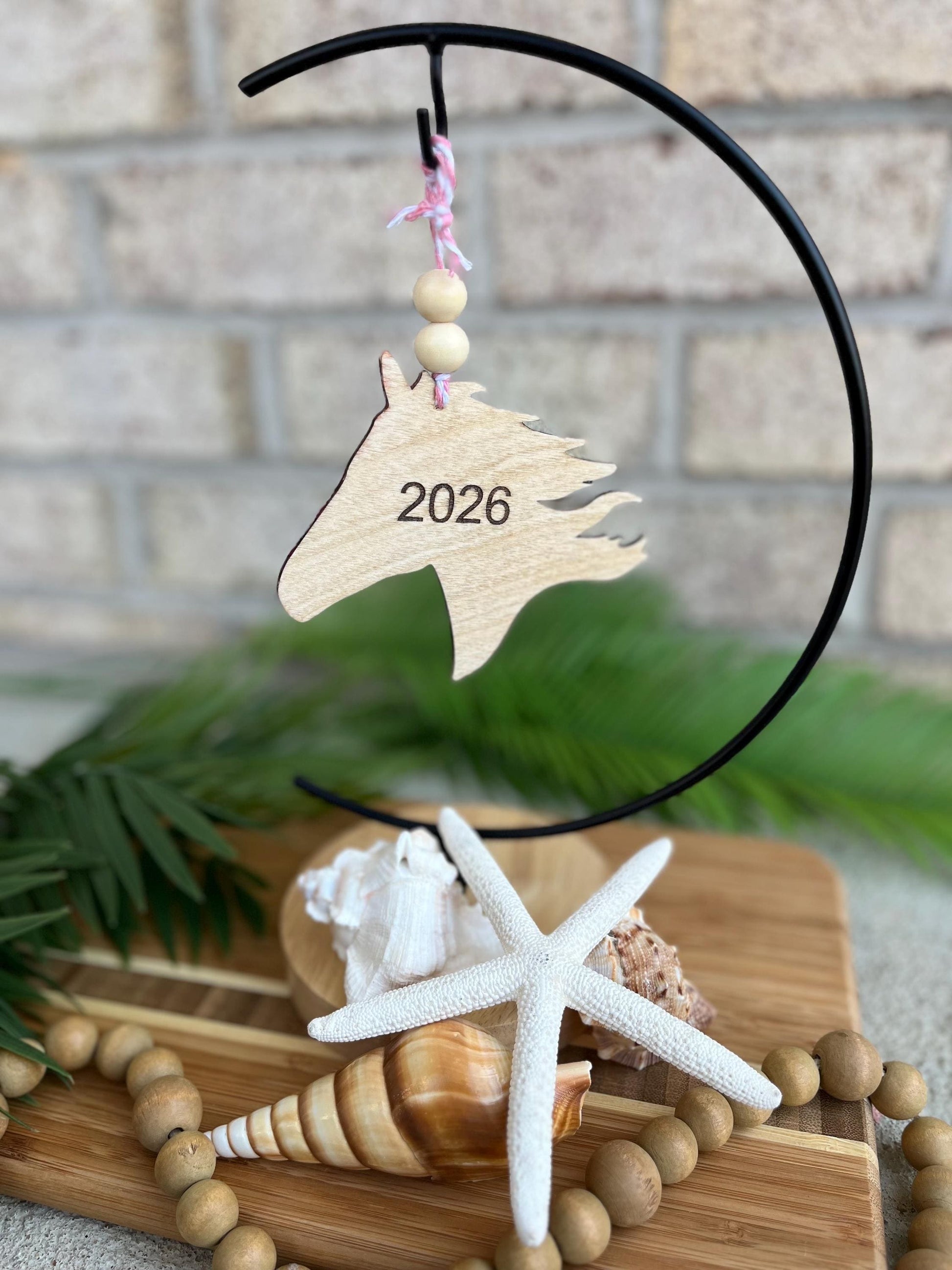 2026 Year of the Horse ornament with 2026 engraved on the back hanging on an ornament hook amongst seashells and palm leaves.
