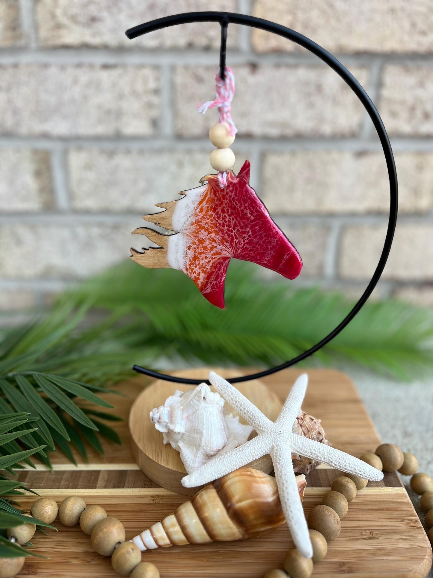 Year of the Fire Horse 2026 ornament with red, orange, and white wave pattern hanging on an ornament hook among seashells and palm leaves.