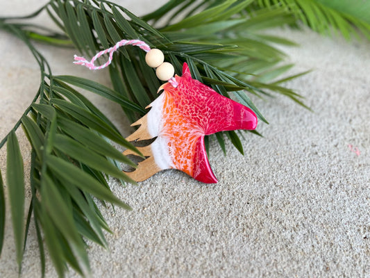 May include: A decorative ornament in the shape of a horse head. The ornament features a vibrant color scheme of red, orange, and white, with a glossy finish. It is adorned with wooden beads and a pink string, and is set against a backdrop of green leaves.