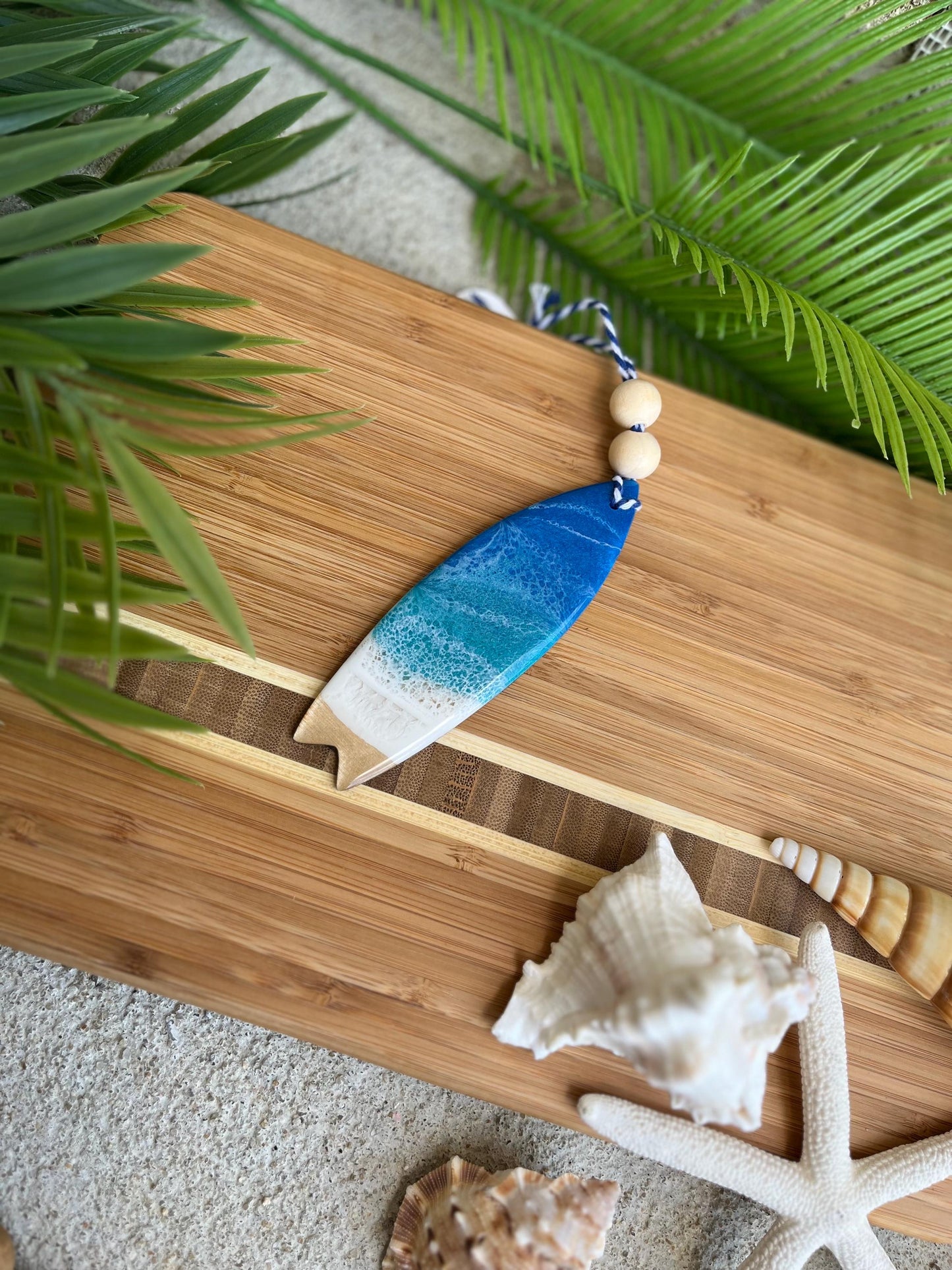 Ocean Resin Art Surfboard Ornament with blue and white waves laying against a bamboo board with palm leaves and seashells