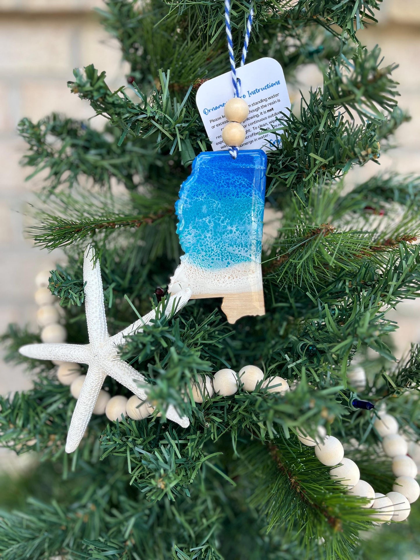 Mississippi ornament featuring ocean resin art and finished with two wooden beads and blue and white twine. Hanging on a Christmas tree with a wooden garland and starfish.
