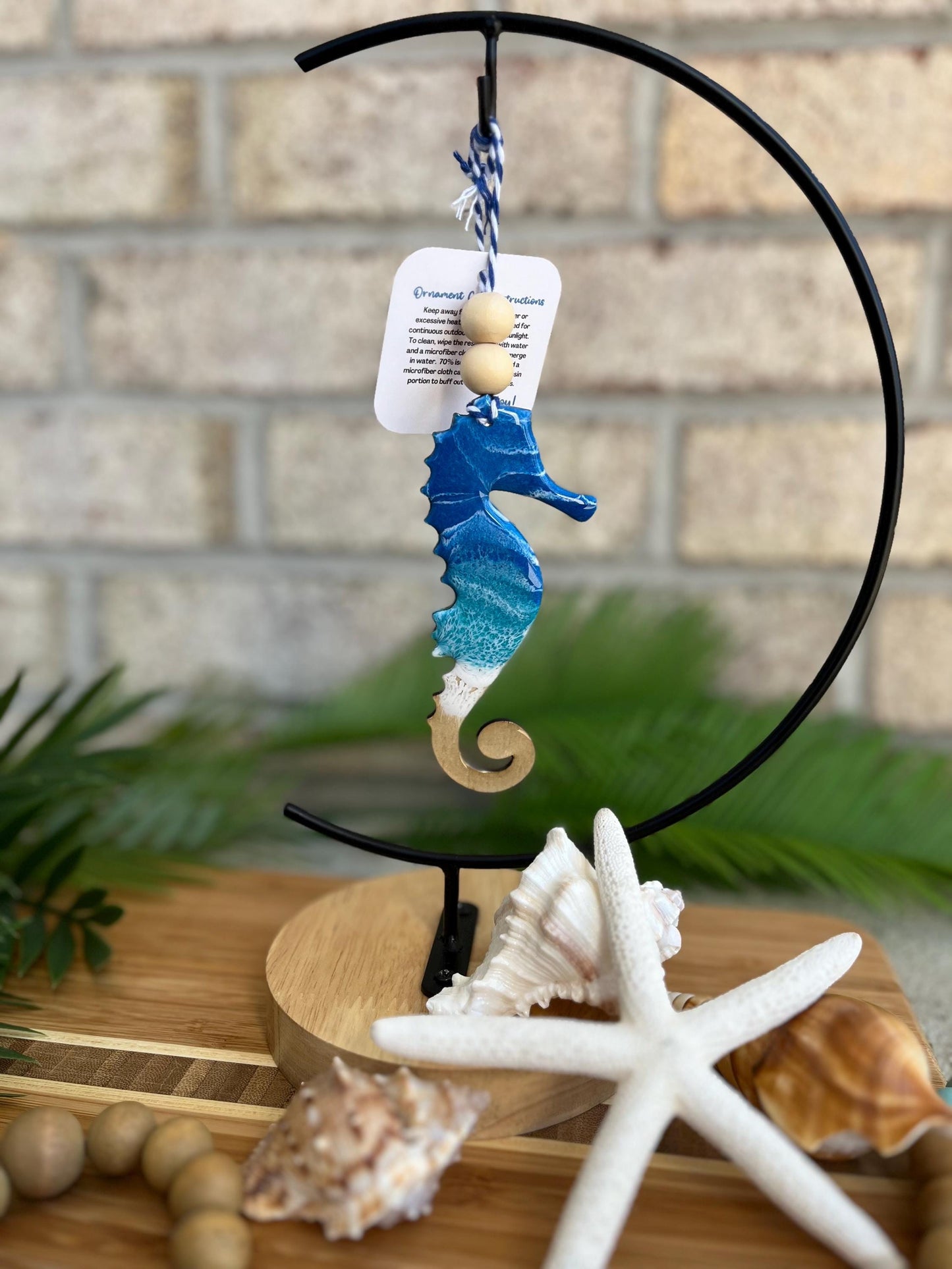 Seahorse ornament with ocean resin art and finished with two wooden beads and blue and white twine. Hanging on an ornament hanger among seashells and a bamboo board.