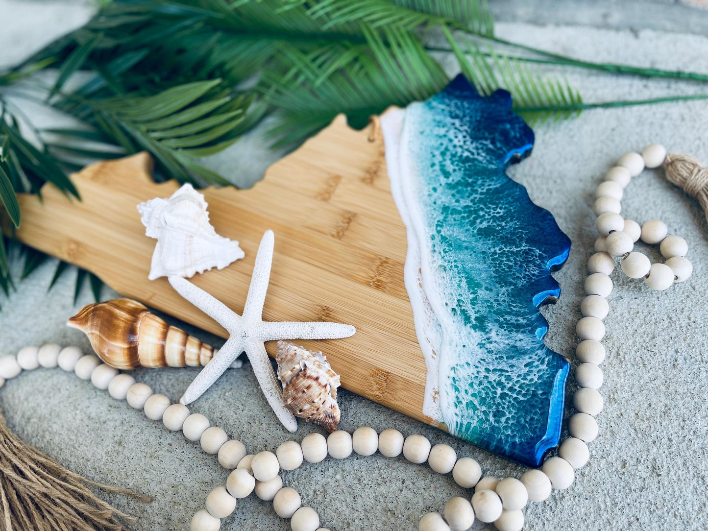 Virginia shaped bamboo charcuterie board with blue and teal ocean resin waves along the coastline portion of the board. Laying against palm leaves and seashells.