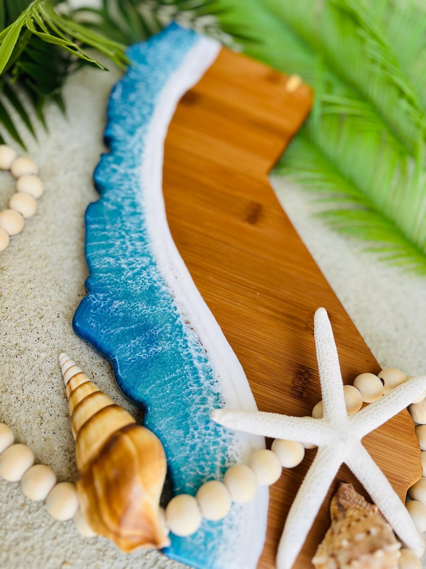 California shaped bamboo charcuterie board with blue and teal ocean resin waves along the coastline portion of the board, laying against palm leaves and seashells.