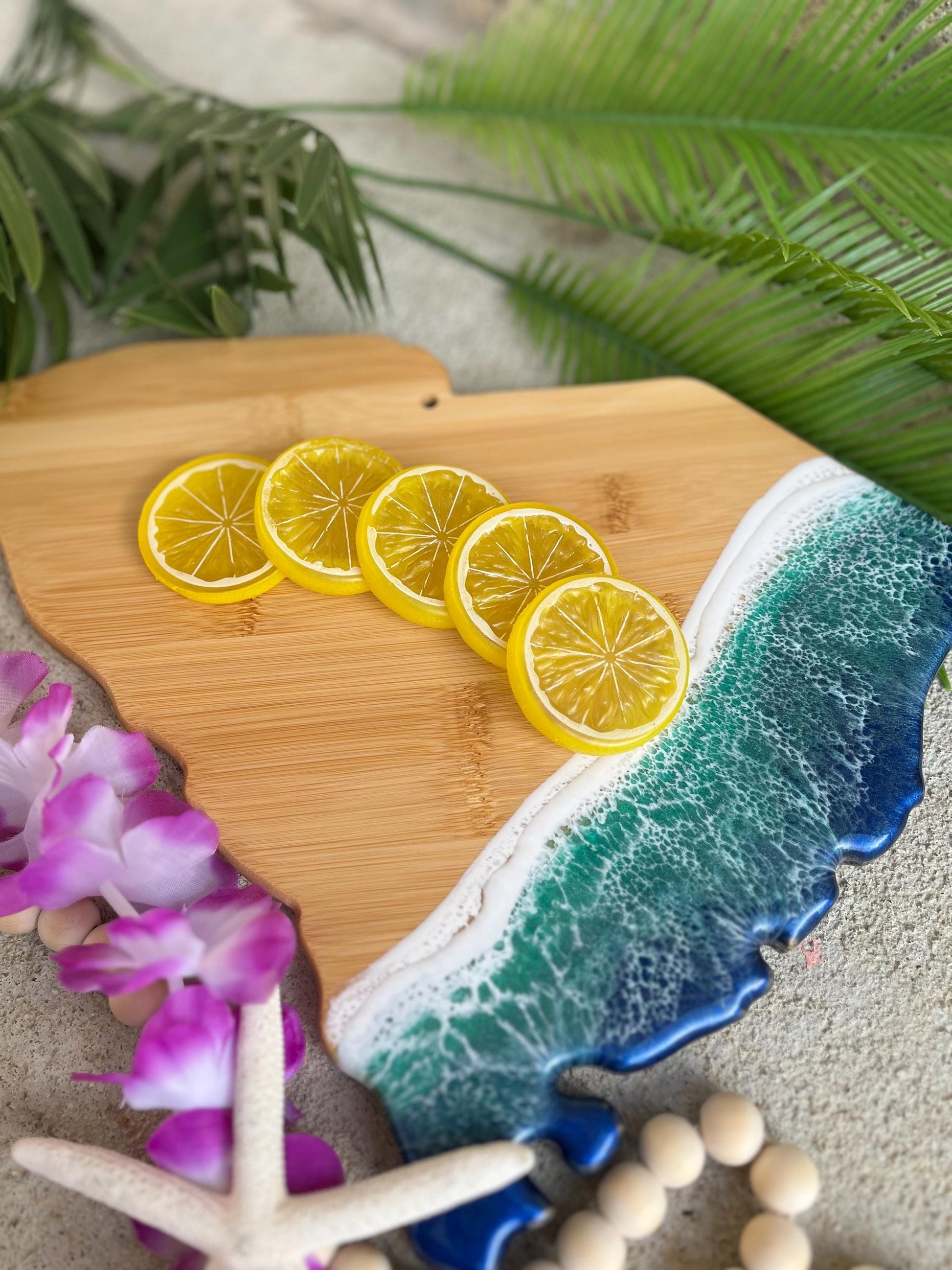 a South Carolina shaped bamboo serving board with blue and green ocean resin art on the coastal side. Shown with lemon slices and laying against palm leaves and seashells.