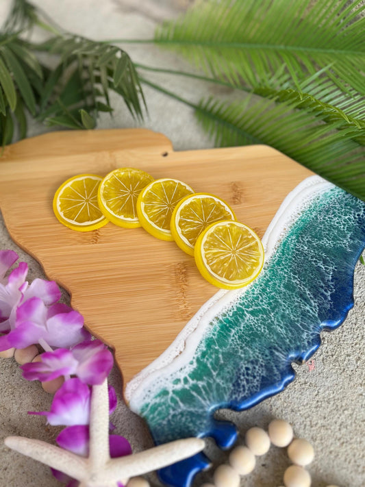 a South Carolina shaped bamboo serving board with blue and green ocean resin art on the coastal side. Shown with lemon slices and laying against palm leaves and seashells.