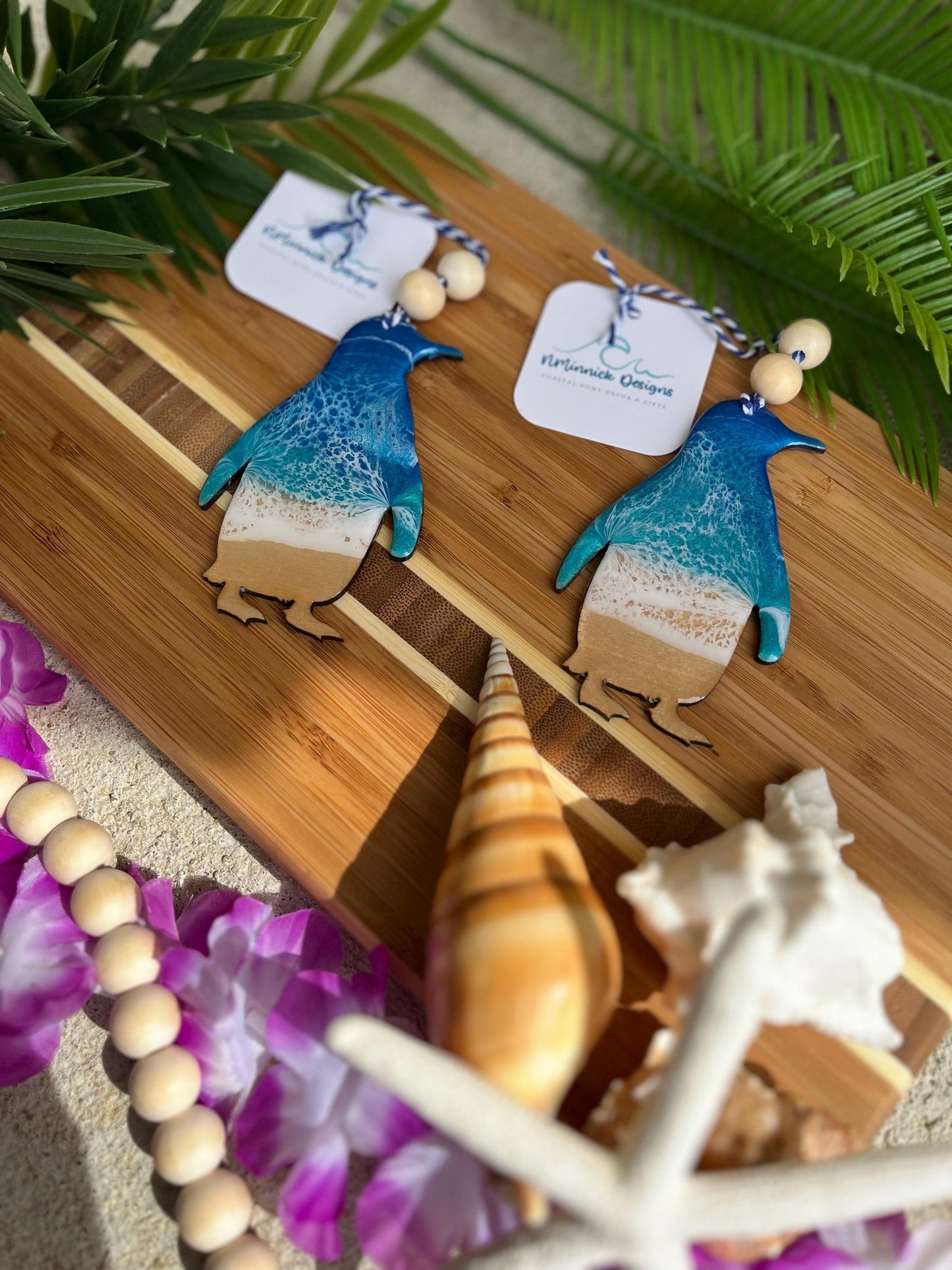 Two penguin ocean resin ornaments laying on a bamboo board among seashells and palm leaves
