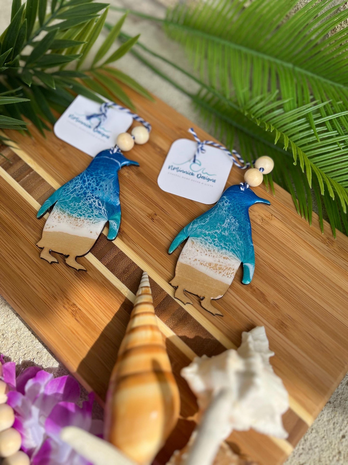 Two penguin ocean resin ornaments laying on a bamboo board among seashells and palm leaves