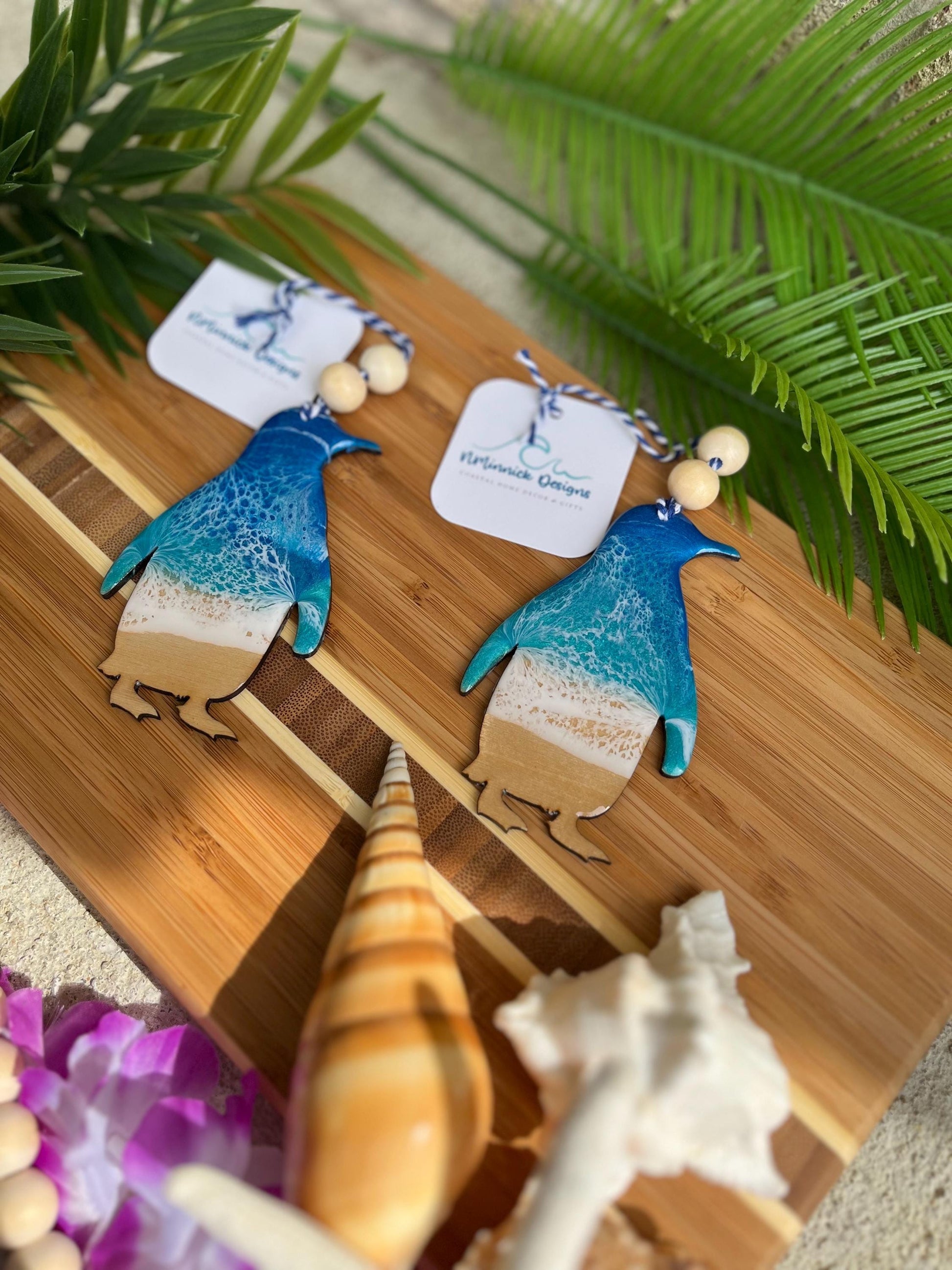 Two penguin ocean resin ornaments laying on a bamboo board among seashells and palm leaves