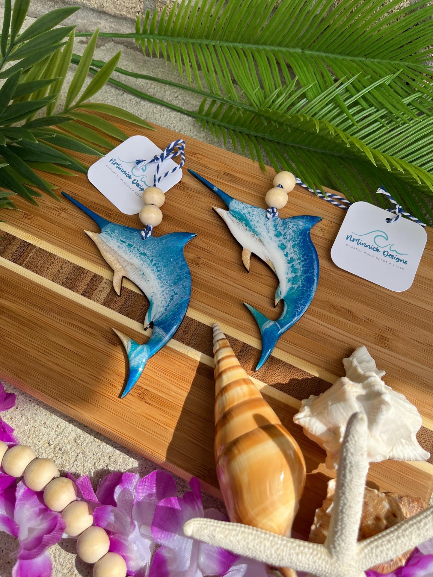 Two Marlin Shaped Ocean Ornaments laying on a bamboo board among sea shells and palm leaves