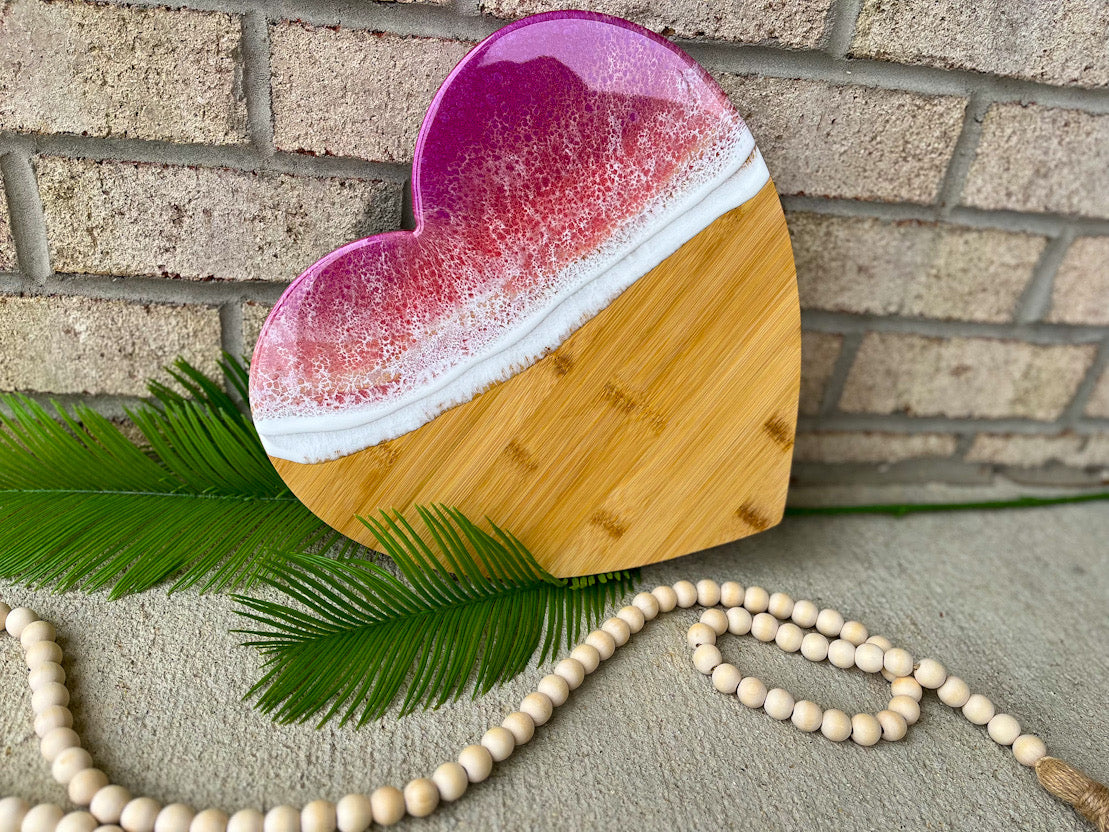 heart shaped charcuterie board with magenta, coral, and golden colored ocean resin waves on top. Setting against a white brick background with palm leaves and wooden beads.