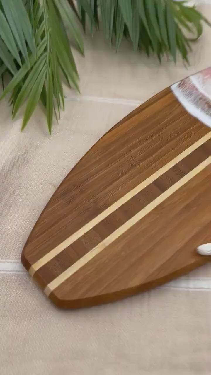 Surfboard-shaped bamboo cutting board with the top third of the board covered in a coral pink and gold tinted ocean resin art resembling the waves of the ocean. Laying on a tan beach towel next to a natural starfish (not included).