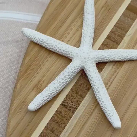 Surfboard-shaped bamboo charcuterie board. Top portion covered in blue and teal ocean resin art resembling the waves of the ocean. Laying on a tan beach towel next to a natural starfish (not included) and adorned with three orange slices and a matching cheese knife (not included).