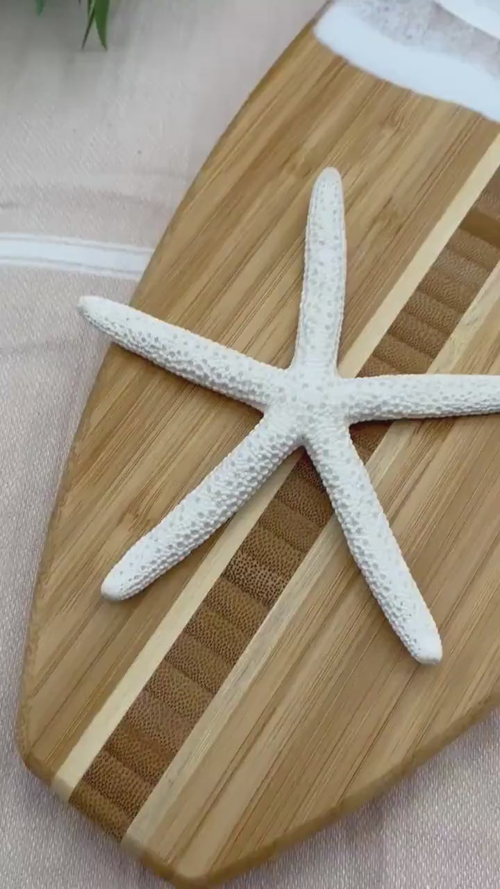 Surfboard-shaped bamboo charcuterie board. Top portion covered in blue and teal ocean resin art resembling the waves of the ocean. Laying on a tan beach towel next to a natural starfish (not included) and adorned with three orange slices and a matching cheese knife (not included).