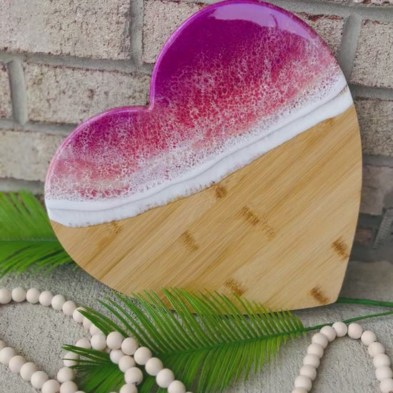 heart shaped charcuterie board with magenta, coral, and golden colored ocean resin waves on top. Setting against a white brick background with palm leaves and wooden beads.