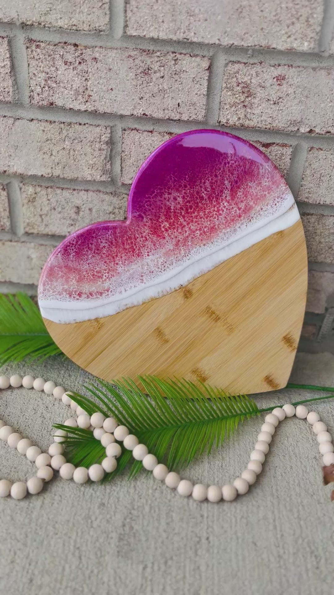 heart shaped charcuterie board with magenta, coral, and golden colored ocean resin waves on top. Setting against a white brick background with palm leaves and wooden beads.